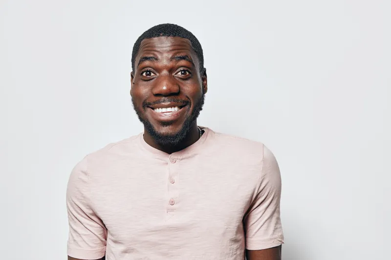 African American man with dark skin smiling and looking at camera wearing light pink shirt on plain background. Portrait, happy expression and casual style with short hair and beard.