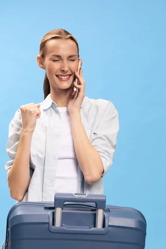 Happy young woman with suitcase talks on phone and clenches fist in excitement against blue background. Concept of travel, vacation, communication, and positive emotions.