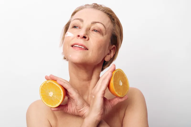 Mature woman with natural skin applies cream and holds two orange halves in hands, posing on white background. Concept for skincare, healthy lifestyle, vitamin c, and beauty care.