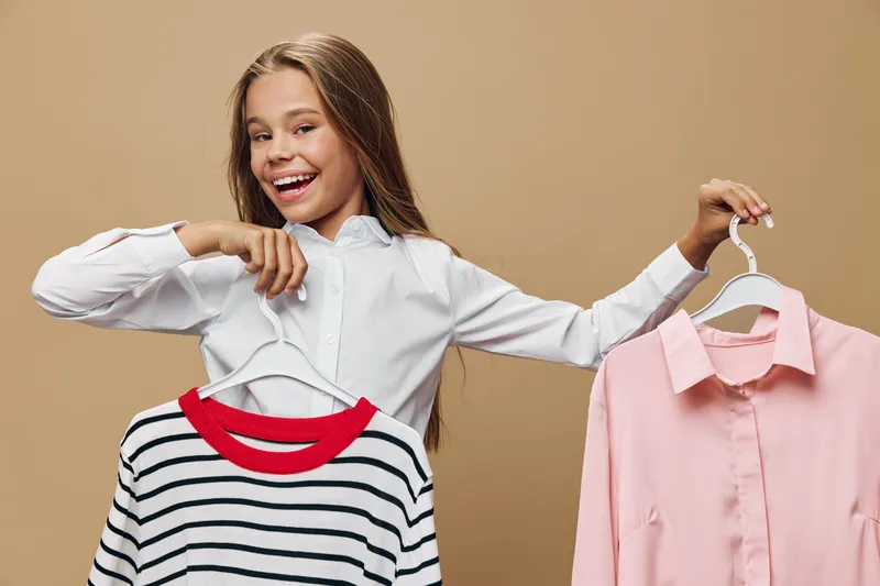 Girl smiling and holding two clothes hangers with striped sweater and pink shirt on beige background. Fashion, outfit choice and casual style for young female with long hair indoors.