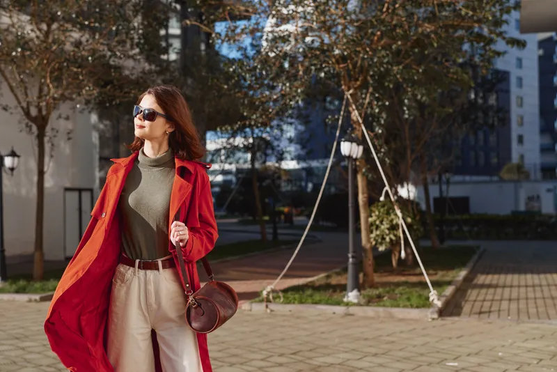 A stylish woman in a red cape strolls through a sunlit urban plaza, wearing a green turtleneck and beige trousers, carrying a brown bag. Concept: chic street style and confident urban fashion.