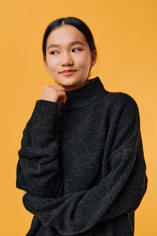 Young woman wearing black turtleneck sweater poses looking to side with thoughtful expression on orange background. Female model in casual clothing with natural makeup and hairstyle indoors.