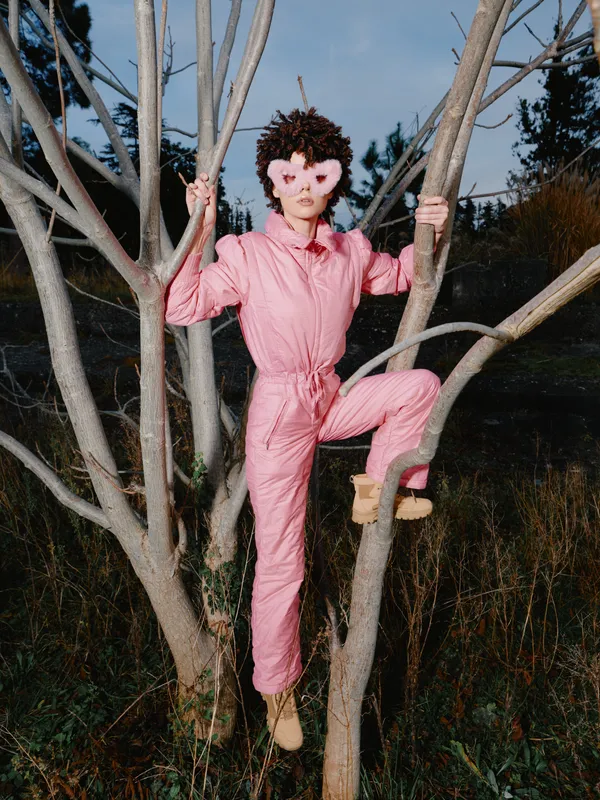 Person in a pink jumpsuit climbing a tree in an outdoor setting, wearing a mask with eerie expression, bare branches and field behind under dim light.