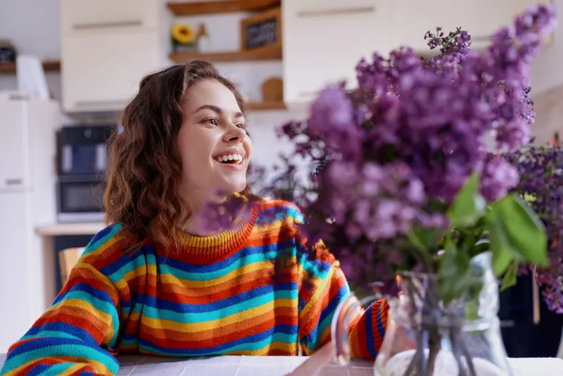 Young woman sits at a bright indoor table wearing a colorful striped sweater, smiling warmly as vibrant flowers bloom nearby, creating a cozy lifestyle moment