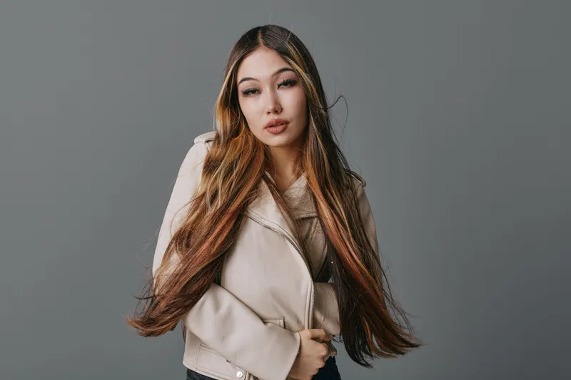Young woman with long hair wearing beige jacket posing confidently against gray background in studio. Fashion portrait, beauty and casual style with natural makeup and modern outfit.