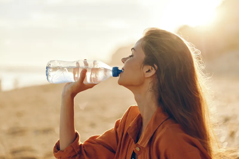 Woman drinking water hydration outdoors beach sunlight health concept, summer vacation on coast, orange shirt female in side profile, holding clear bottle, calm refreshing mood