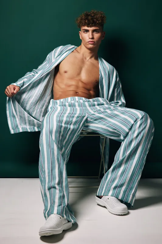Young man with curly hair wearing striped pajamas and white shoes sitting on a white bench against dark green background. Fashion, casual style and relaxed pose in studio.