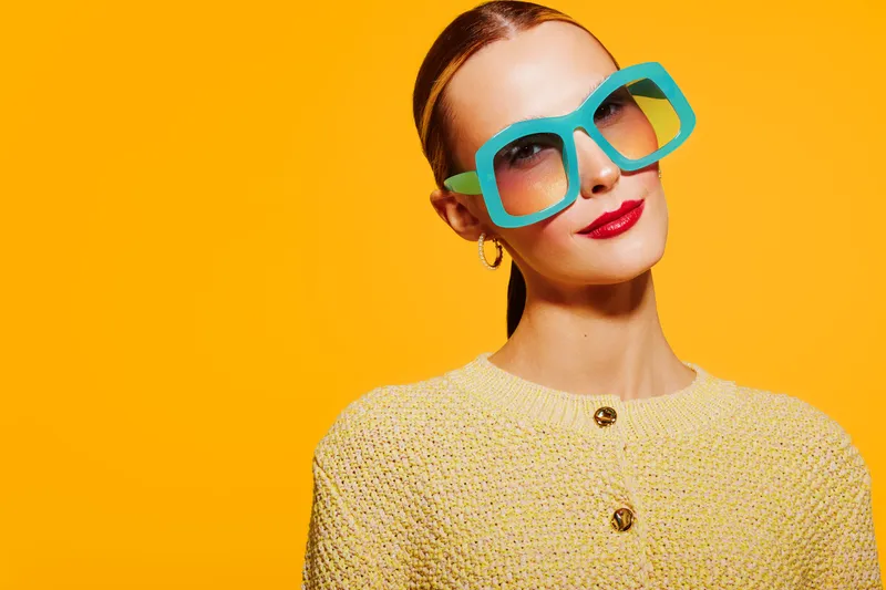 Stylish woman with blue oversized sunglasses and red lipstick poses against bright yellow background. Fashion portrait with vibrant colors and confident expression. Concept of summer style.
