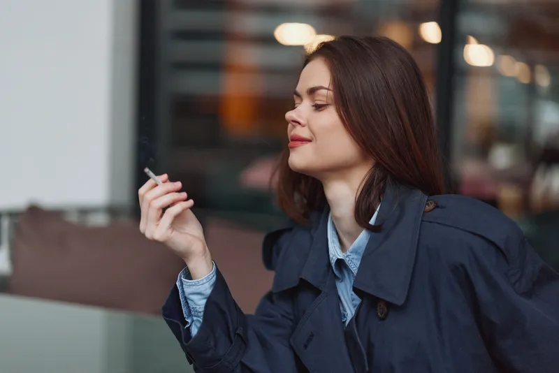 Young woman with long brown hair smoking cigarette outdoors near glass wall. Casual outfit with navy coat and blue shirt. Urban lifestyle concept with relaxed mood.
