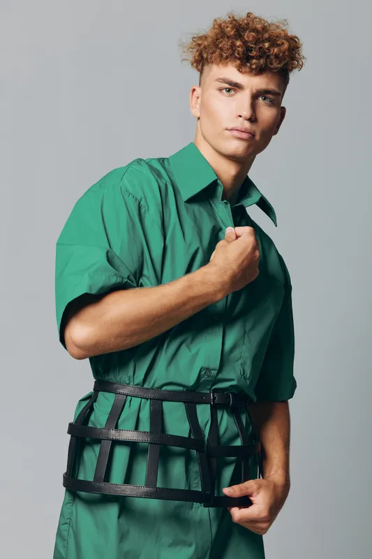 Young man in green shirt posing with serious expression and curly hair studio fashion contemporary style wearing harness accessory over outfit on gray background.