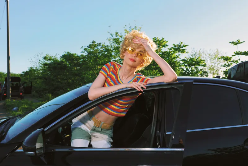 Cheerful woman outdoors beside a sleek car, striped top and casual denim, sunlit moment with retro film color, relaxed pose under a bright sky and lush greenery colors