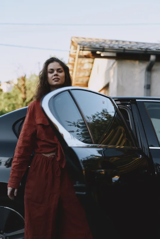 A woman in a red coat stands beside a car outdoors, capturing a modern lifestyle moment in an urban street scene. Casual, confident, stylish, and ready for the day