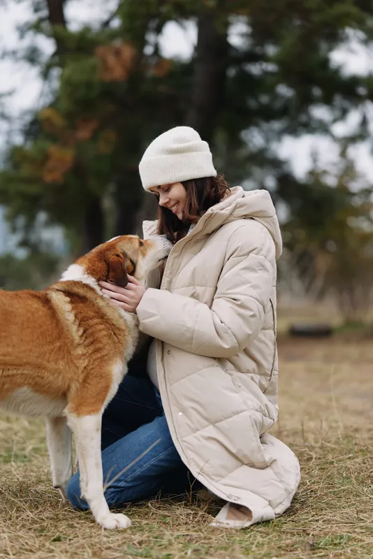 Woman kneeling in a light coat and beanie, cuddling a friendly dog on a grassy field, autumn light and candid moment of companionship in an outdoor park setting