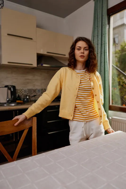 Young woman with curly hair stands in a bright kitchen wearing a striped orange shirt and yellow cardigan, relaxed at a table with natural light and a cozy home atmosphere.
