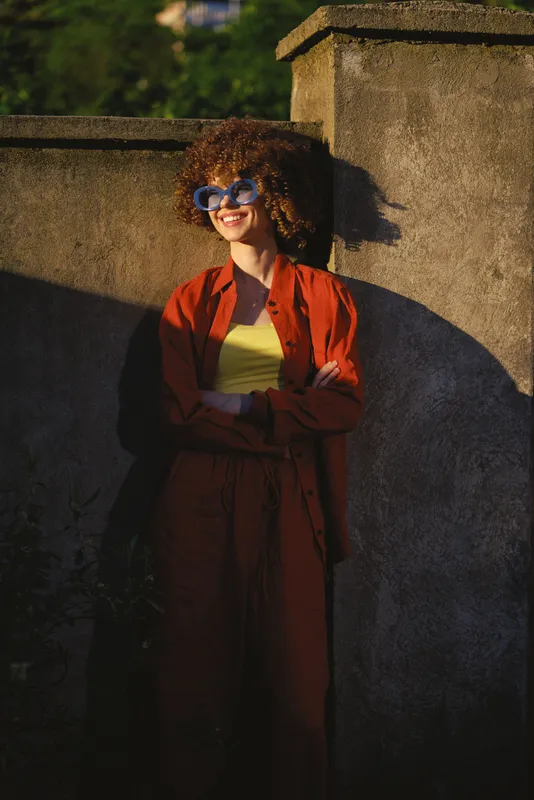 A young woman with curly hair leans against a stone wall outdoors, wearing a red coat and yellow top, smiling behind sunglasses in bright daylight