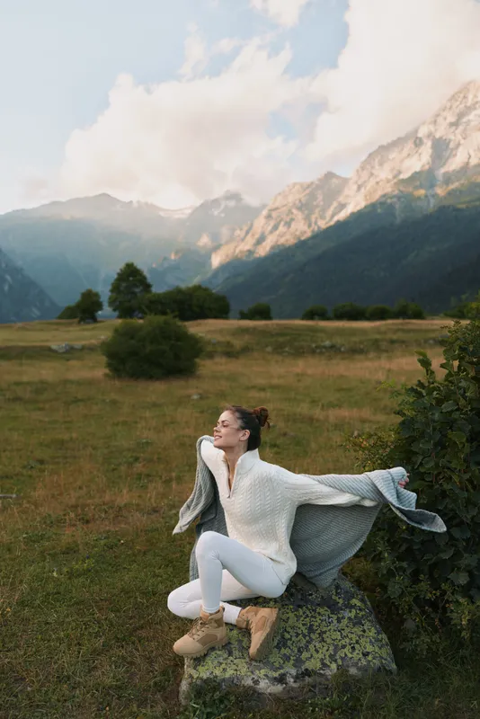 A woman sits on a rock in a white knit sweater and boots, stretching her arms, gazing toward distant mountains, a calm outdoor lifestyle moment in nature.