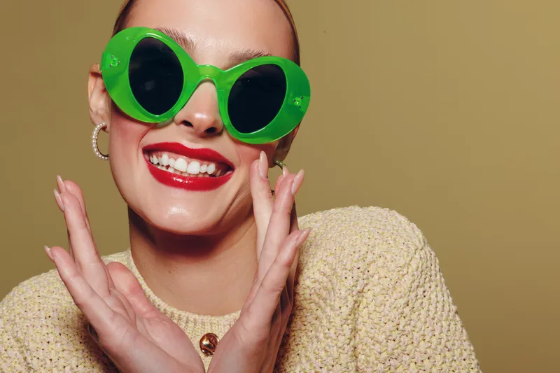 Smiling woman with bright red lipstick wears large green sunglasses and beige sweater. Close-up portrait on neutral background. Concept of fashion, style, and joyful expression.