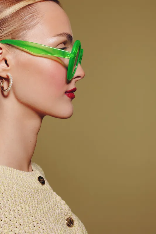 Profile of a stylish woman wearing green transparent sunglasses and gold hoop earrings. She has red lipstick and a beige knitted sweater with buttons on a neutral background.