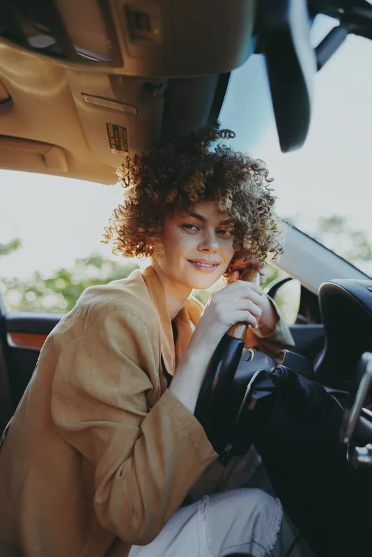 Casual woman with curly hair sits in car, hands on steering wheel, relaxed lifestyle scene, warm retro film color, daylight interior shot, candid smile, urban commute moment