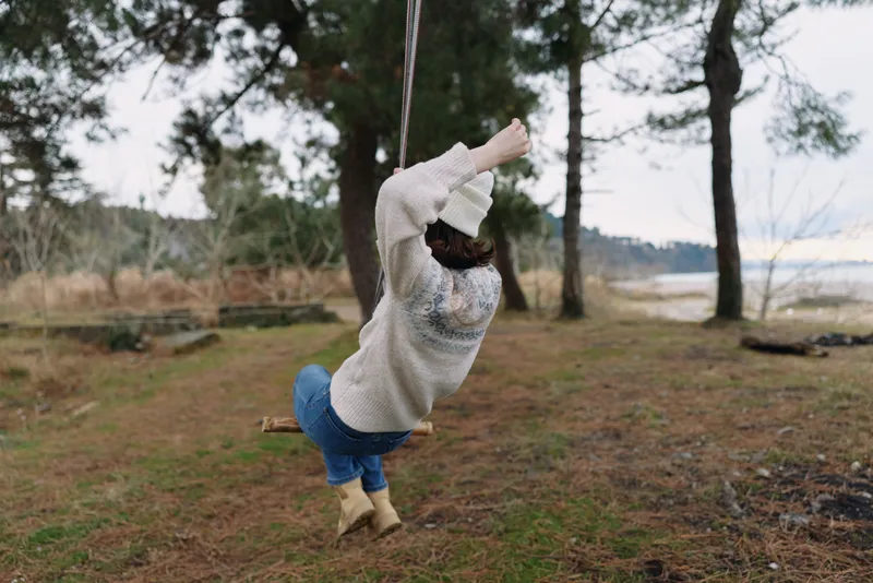 Child on a rope swing in a park by a lake, wearing a beige sweater and blue jeans, twirling gently as wind rustles trees, capturing carefree outdoor play.