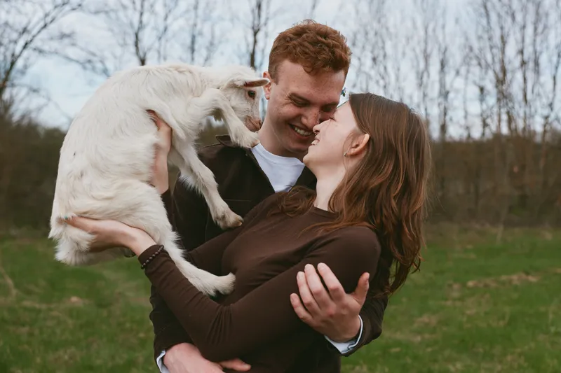 Joyful couple holding a playful goat kid in an open field, celebrating rural life. Natural daylight, candid affection, bonding, outdoor leisure, farm companionship, warm, relaxed mood.