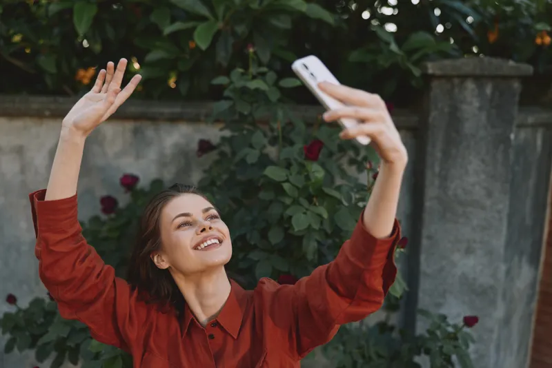 Young woman takes a cheerful selfie in a sunlit garden, smiling with raised arms and a red shirt, capturing a candid moment outdoors, radiating zest, warmth, and relaxed confidence