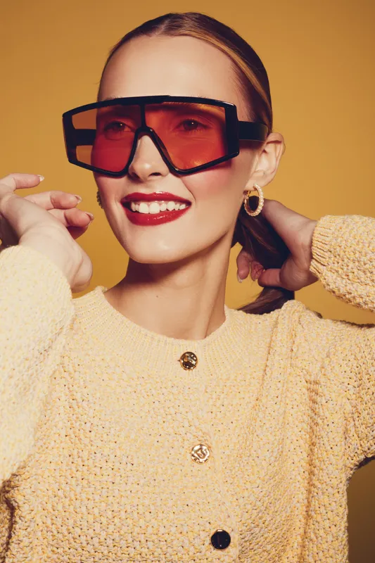 Smiling woman with red sunglasses and red lipstick wears yellow knitted sweater, posing with hands behind head on yellow background. Concept of fashion, style, and confident beauty.