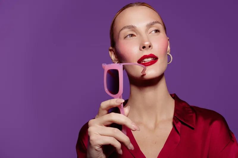 Stylish woman with red lipstick and gold earrings holds pink sunglasses near face on purple background. Fashion portrait with vibrant colors and confident expression in studio lighting.