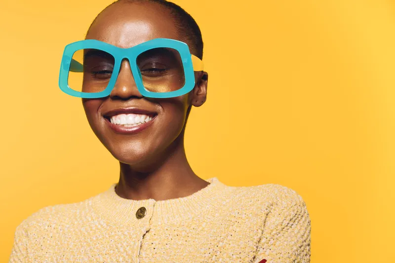 Happy African woman with dark skin wearing oversized blue sunglasses and yellow sweater smiles with closed eyes on bright yellow background. Concept of joy, fashion, and summer style.
