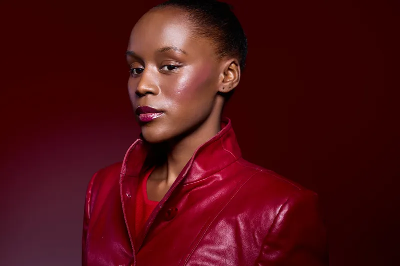 Confident African woman with dark skin wearing a red leather jacket poses against a deep red background. Studio portrait highlights fashion, beauty, and elegance concepts.