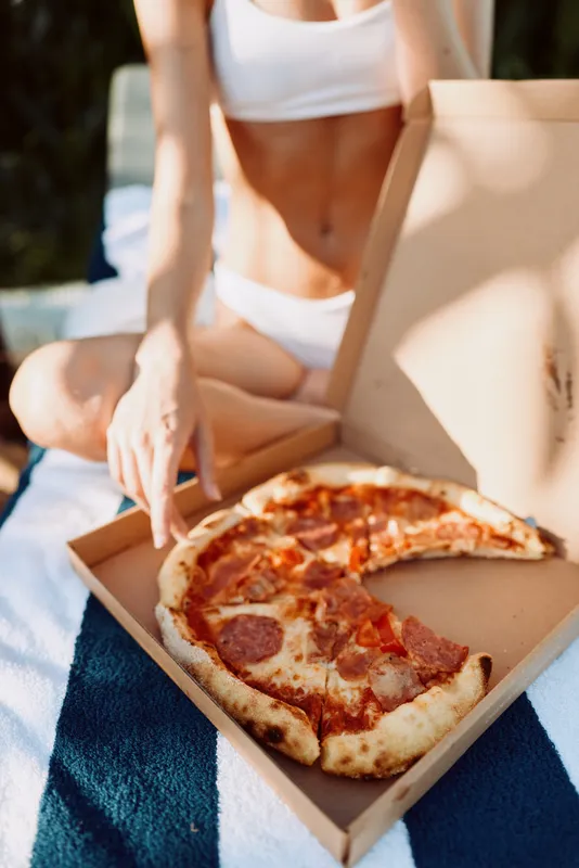 Casual outdoor scene shows a woman in white attire seated on a blanket with an open pizza box, reaching for a slice, conveying a relaxed lifestyle, sunny mood, and summertime leisure.