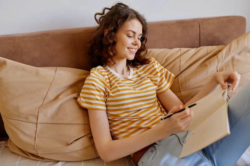 A young woman with curly hair sits on a beige sofa in a bright living room, writing on a clipboard with a relaxed, home lifestyle vibe.