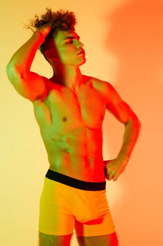 Young man posing with hand on head in colorful lighting wearing boxer shorts. Studio workout portrait showcasing fit body and confident muscle definition with vivid background colors.