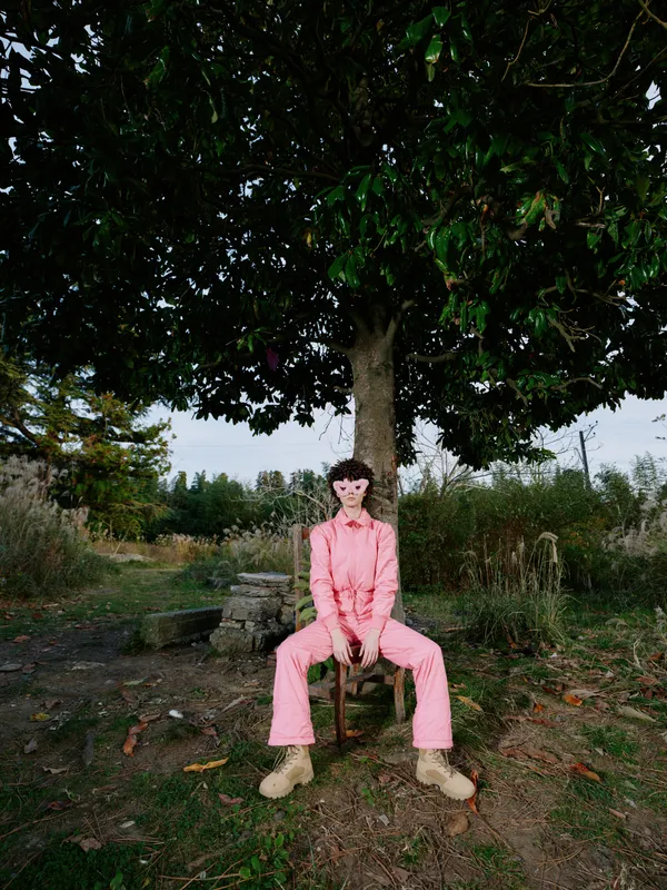 Outdoor portrait of a solo model in a pink jumpsuit seated under a large tree in a rural setting, showcasing natural light, calm mood, and casual pose amid fields and rustic surroundings.