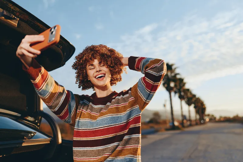 Woman lifestyle outdoor, with curly hair and smile, car washing, holding a hose to rinse the front bumper on a driveway, warm sunset light, summer sky