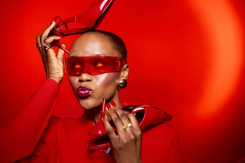 Stylish African woman with dark skin poses with red high heels and matching sunglasses on red background. Fashion concept with bold accessories and confident attitude.
