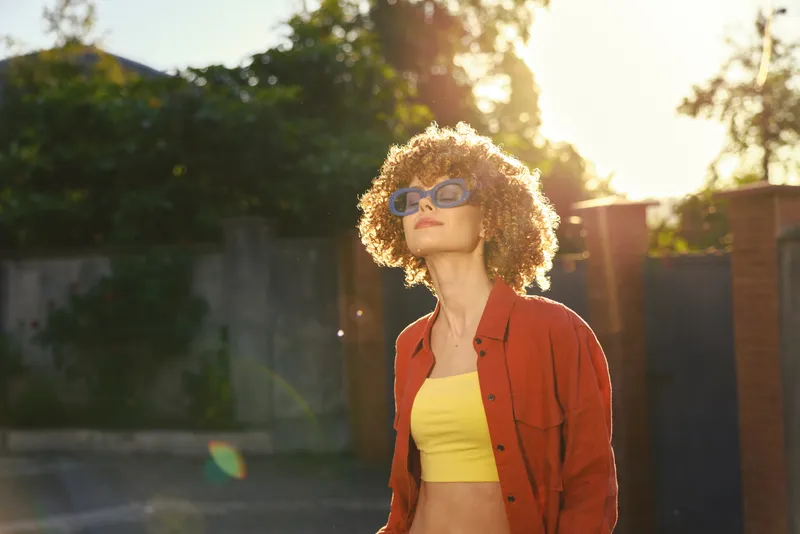 Woman lifestyle portrait outdoors, curly haired woman in a yellow crop top and red shirt, sunny glow, casual mood, blue sunglasses, vibrant warm street scene