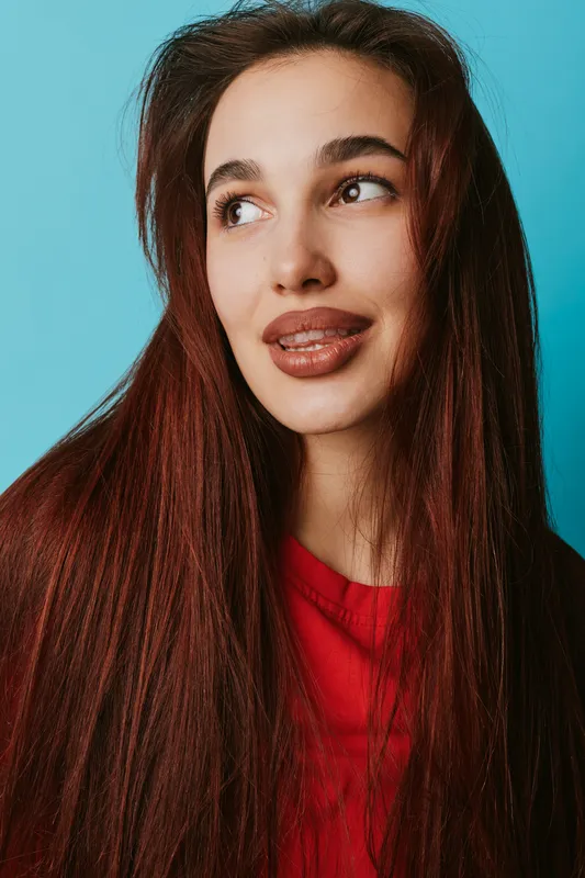 Young woman with long brown hair and natural makeup wearing red sweater looking sideways with slight smile against blue background in studio portrait photography.