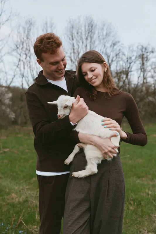 Young couple holding a small white goat outdoors in a green field during early spring, enjoying a tender moment together in casual warm clothing.