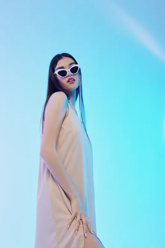 Young woman in a beige dress and white sunglasses poses confidently against a blue gradient background. Concept of woman beauty fashion and modern style.