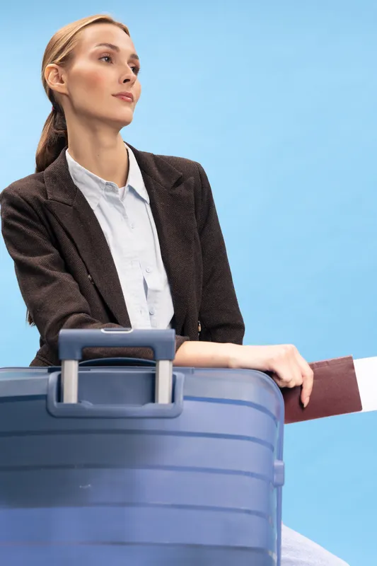 Confident businesswoman in blazer with suitcase and passport waits for flight against blue background. Concept of travel, business trip, and airport waiting time.