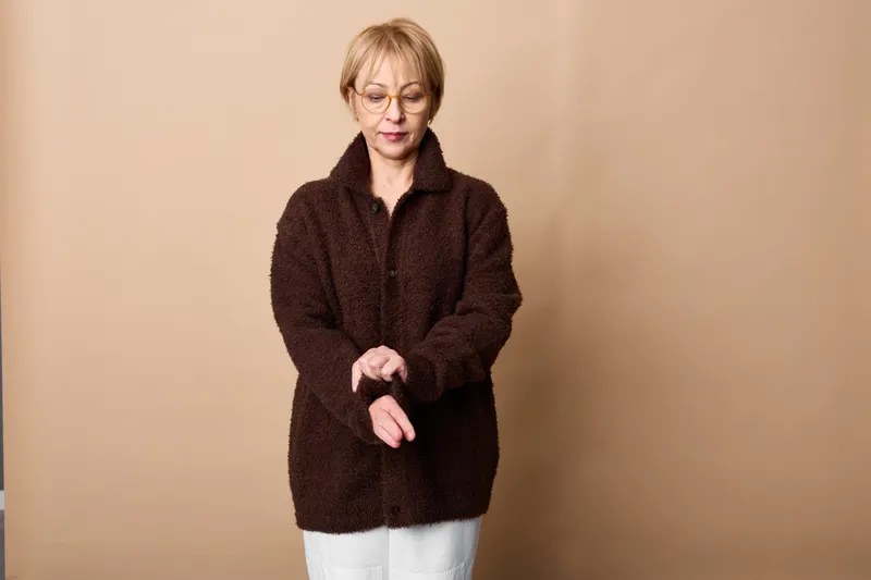 Elderly woman in dark sweater on beige background, hands held together, looking down with tired expression, concept of loneliness and aging, quiet sadness
