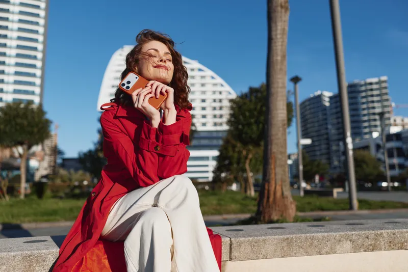 A young woman in a red coat sits outdoors on a sunny day, holding a smartphone and smiling with closed eyes, enjoying a peaceful moment in an urban park.