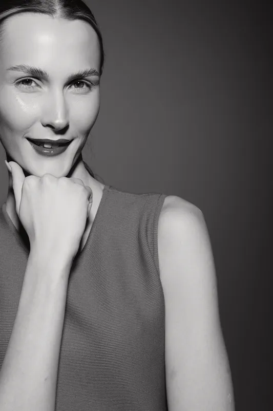 Close-up portrait of a smiling Caucasian woman with light skin touching chin. Studio black and white photography with soft lighting highlights facial features and expression.