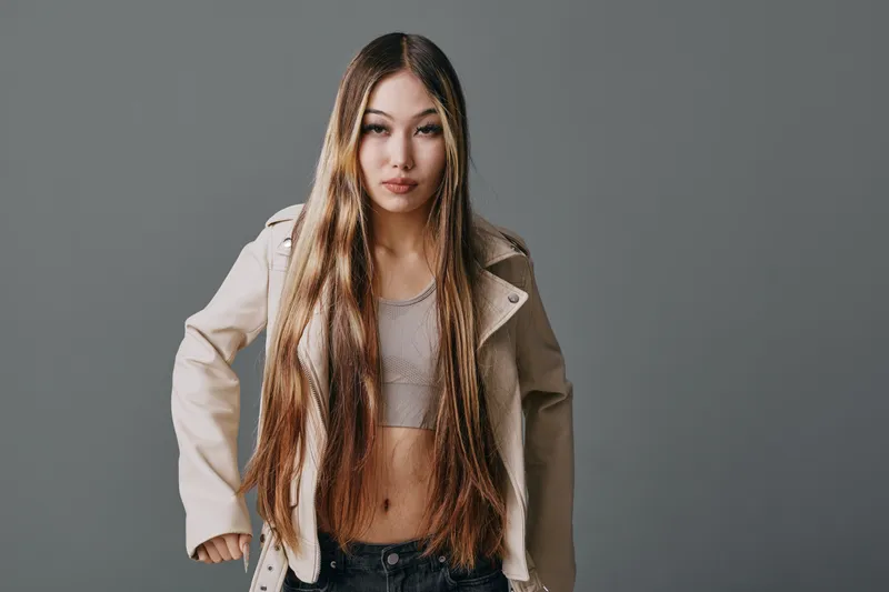 Young woman with long hair wearing a cropped top and light jacket posing confidently against gray background. Casual style, fashion, beauty and portrait with natural makeup in studio.