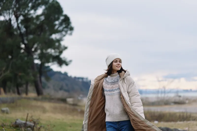 Woman in a warm coat and knitted scarf walks outdoors by a lake, wearing a beanie. Windy landscape, calm sky, and casual winter fashion in a natural setting.