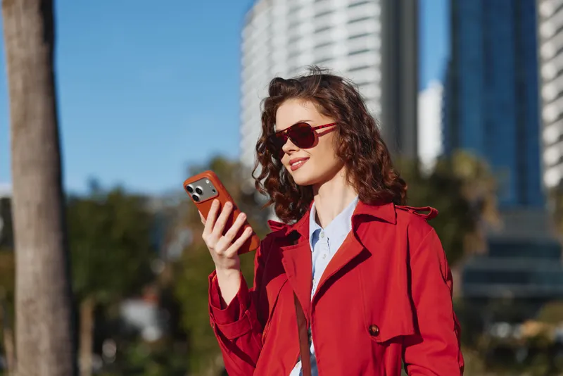 Young woman in a red trench coat outdoors, smiling subtly as she looks at her phone, urban daylight, sunglasses on, cityscape blur behind.