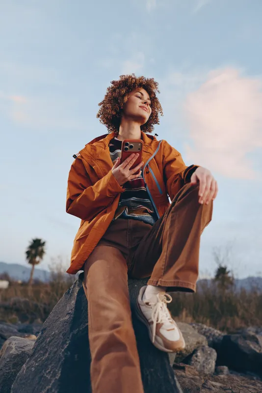 Woman lifestyle outdoors portrait, curly hair, orange jacket, holding smartphone, looking at phone, sitting on rock in mountain landscape, clouds and sky, warm sunset daylight, smile