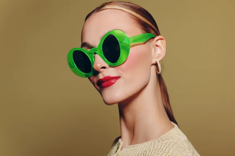 Stylish woman with slicked back hair wearing large green sunglasses and red lipstick poses against beige background. Concept of fashion, beauty, accessories, and confident attitude.