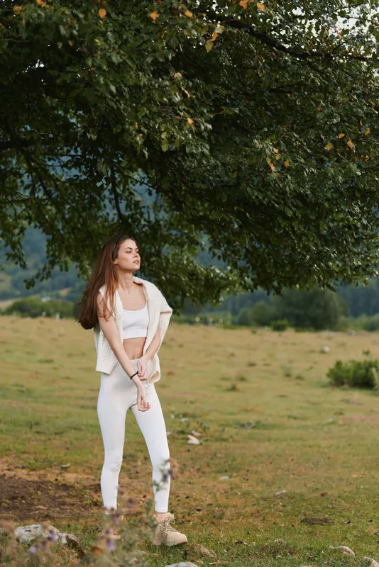 Young woman in white athleisure stands alone in a sunlit field beneath a large leafy tree, gazing into the distance with a calm, introspective mood as a soft breeze moves her hair and the golden hour
